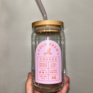 May include: A clear glass jar with a bamboo lid and a pink and orange label that reads "COLD BREW COFFEE" with the text "FRESHLY BREWED", "ROAST", "EST. EVERY MORNING", and "NET WT. 16 oz".
