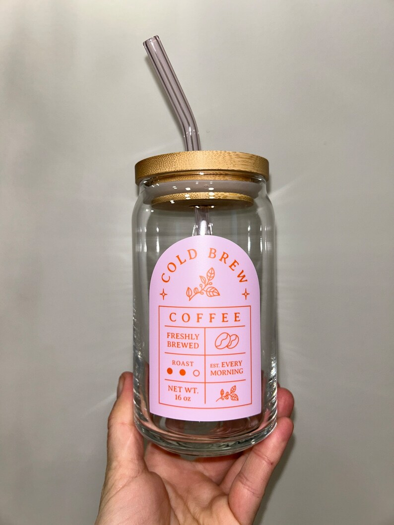 May include: A clear glass jar with a bamboo lid and a pink and orange label that reads "COLD BREW COFFEE" with the text "FRESHLY BREWED", "ROAST", "EST. EVERY MORNING", and "NET WT. 16 oz".