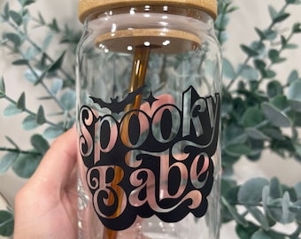 May include: A clear glass tumbler with a bamboo lid and a gold-colored straw. The tumbler features the words "Spooky Babe" in a black and silver design, with a bat graphic. The glass is held by a hand.