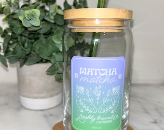 Puede incluir: Un tarro de vidrio transparente con tapa de bambú y una pajita de vidrio verde. El tarro tiene una etiqueta que dice "MATCHA matcha" con un diseño floral y el texto "recién hecho cada mañana".