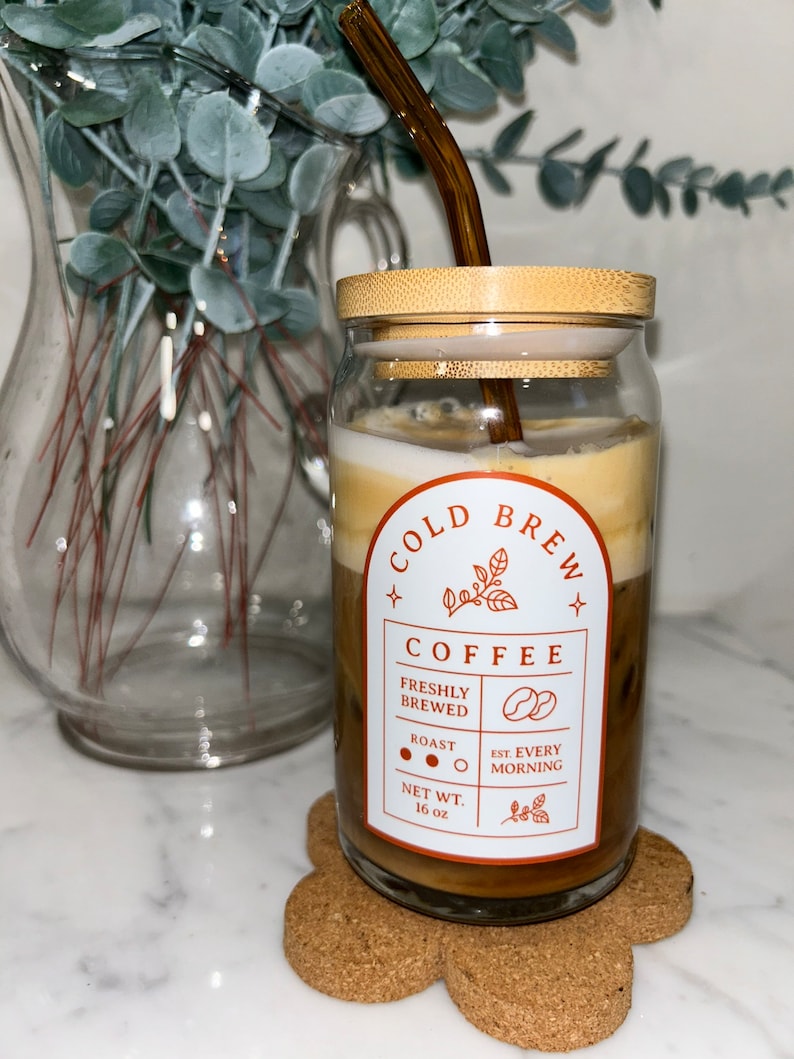 May include: A glass jar with a bamboo lid filled with cold brew coffee. The jar has a label that says "Cold Brew Coffee" with the text "Freshly Brewed", "Roast", "Est. Every Morning", and "Net Wt. 16 oz". A brown glass straw is in the jar.