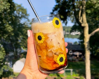 May include: A glass of iced coffee with a metal straw, decorated with sunflower decals. The coffee is a light brown color and has a layer of cream on top. The glass is held by a hand with a gold bracelet.