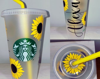May include: A clear plastic reusable cup with a black and green Starbucks logo and a sunflower design. The cup has a yellow straw and the name "Alexa" written on the side.