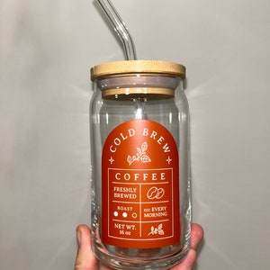 May include: A glass jar with a bamboo lid and a glass straw. The jar has a label that says "Cold Brew Coffee" with the text "Freshly Brewed", "Roast", "Est. Every Morning", and "Net Wt. 16 oz".