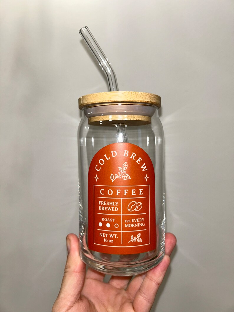 May include: A glass jar with a bamboo lid and a glass straw. The jar has a label that says "Cold Brew Coffee" with the text "Freshly Brewed", "Roast", "Est. Every Morning", and "Net Wt. 16 oz".