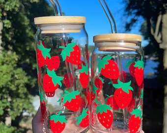May include: Two glass drinking glasses with bamboo lids and clear glass straws. The glasses are decorated with red and green strawberry designs.