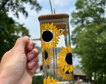 May include: A clear glass mug with a handle and a bamboo lid. The mug has a sunflower design with yellow petals and black centers. A clear drinking straw is inserted through the lid.