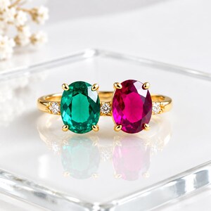 May include: A gold ring featuring two oval-cut gemstones, one vibrant green and one deep pink, set side-by-side. The band is accented with three small, clear round-cut stones, with one positioned between the main gems and one on each side.