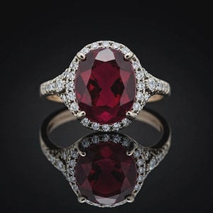 Könnte beinhalten: Ein Ring mit einem oval geschliffenen, tiefroten Edelstein in einer polierten Goldschiene. Der zentrale Stein ist von einem Kranz aus kleinen, runden, klaren weißen Diamanten umgeben, wobei weitere kleine Diamanten in die Ringschiene eingelassen sind.