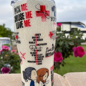 May include: A white tumbler with a glitter finish and a design featuring the characters from the TV show Grey's Anatomy. The tumbler has the text "Pick Me Choose Me Love Me" and "Grey's Anatomy" on it.