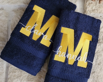May include: Two navy blue towels with large yellow embroidered "M" letters. The towels are personalized with the names "Clara" and "Hannah" in white cursive.