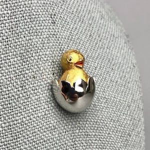 May include: A gold and silver pin of a baby chick hatching from an egg.
