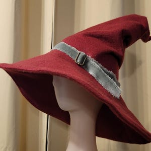 May include: A wide-brimmed, burgundy felt wizard hat with a pointed crown and a gray ribbon with a buckle.