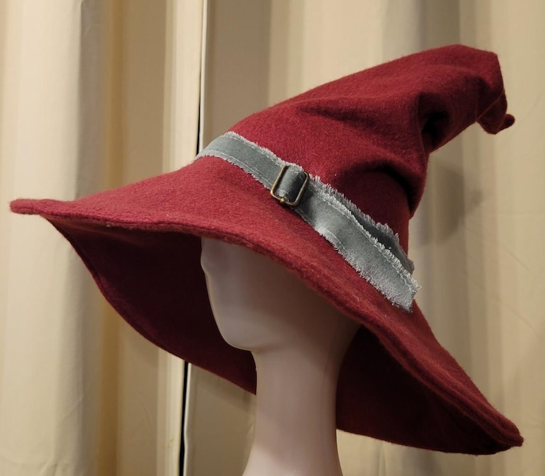 May include: A wide-brimmed, burgundy felt wizard hat with a pointed crown and a gray ribbon with a buckle.