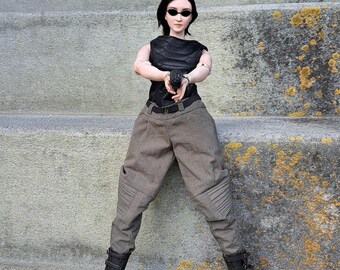 May include: A doll dressed in a black top, gray pants, and black boots, holding a black handgun. The doll is wearing sunglasses and standing on concrete steps. The pants have a pleated design on the knees.