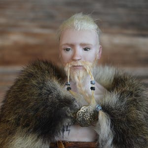 Portrait of a blonde male Viking doll, wearing a long moustache and braided beard and a furr costume. Doll is made of porcelain.