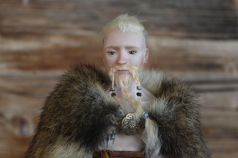 Portrait of a blonde male Viking doll, wearing a long moustache and braided beard and a furr costume. Doll is made of porcelain.