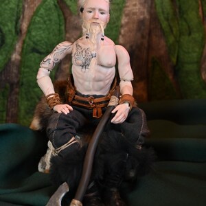 May include: A detailed doll of a Viking warrior, seated with an axe and a staff. The doll has a tattoo of a wolf on its chest and arm. The warrior wears dark pants, fur-lined boots, and a leather belt. The background is a forest scene.