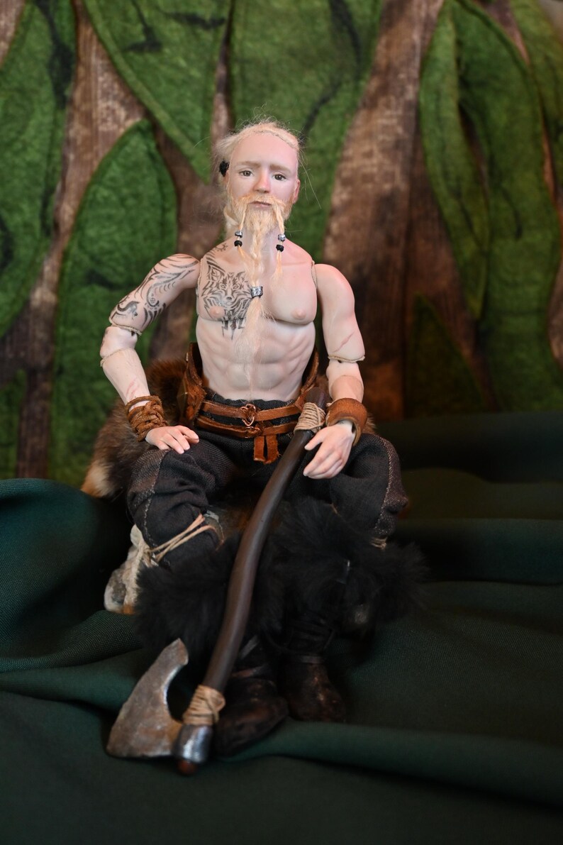 May include: A detailed doll of a Viking warrior, seated with an axe and a staff. The doll has a tattoo of a wolf on its chest and arm. The warrior wears dark pants, fur-lined boots, and a leather belt. The background is a forest scene.