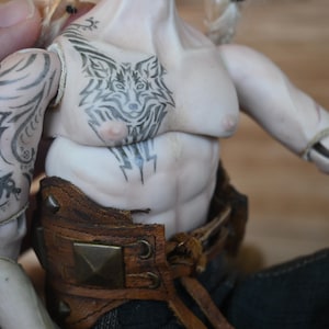 May include: A detailed doll figure with a muscular torso and intricate black tattoos. The tattoos include a wolf design on the chest and patterns on the arm. The doll wears a brown leather belt with metal studs.