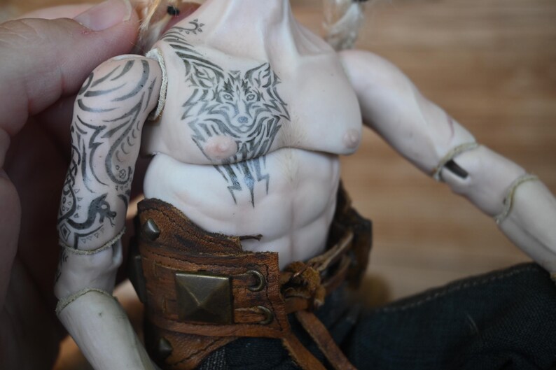 May include: A detailed doll figure with a muscular torso and intricate black tattoos. The tattoos include a wolf design on the chest and patterns on the arm. The doll wears a brown leather belt with metal studs.