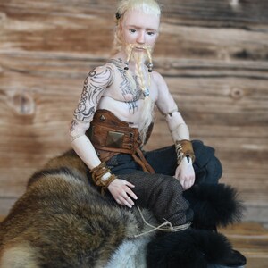 May include: A detailed doll of a Viking warrior with blonde hair and a braided beard, adorned with tattoos and leather accessories. The doll is seated on a fur-covered rock, wearing dark pants and boots.