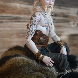 May include: A detailed doll of a male figure with blonde hair and a beard, adorned with tattoos on his arms and chest. He wears a brown leather belt and wristbands, and dark pants. The doll is seated on a fur-like surface.