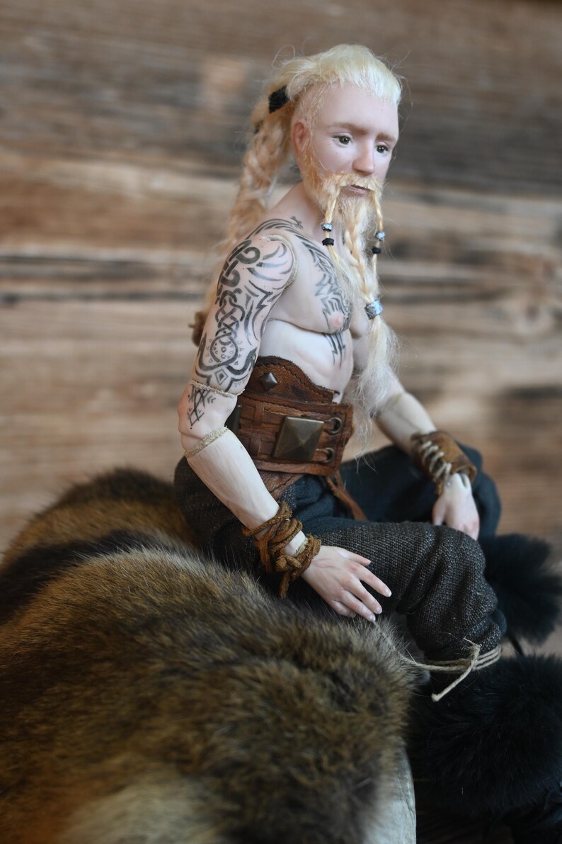 May include: A detailed doll of a male figure with blonde hair and a beard, adorned with tattoos on his arms and chest. He wears a brown leather belt and wristbands, and dark pants. The doll is seated on a fur-like surface.