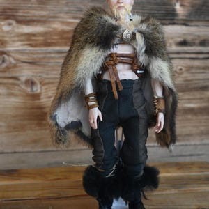 May include: A doll depicting a Viking warrior. The doll wears a fur cape, leather straps, and black pants with fur-trimmed boots. The doll has a blonde beard and hair, and is standing on a white base.