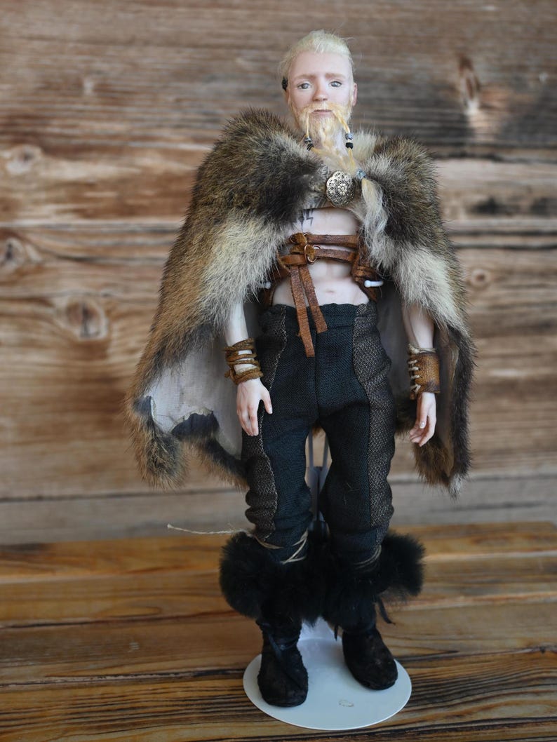May include: A doll depicting a Viking warrior. The doll wears a fur cape, leather straps, and black pants with fur-trimmed boots. The doll has a blonde beard and hair, and is standing on a white base.