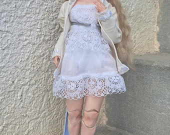May include: A detailed doll with long blonde hair, wearing a white lace dress, a cream-colored jacket, and a silver belt. The doll is standing on a light blue stand. The doll's skin tone is fair, and it has detailed facial features.