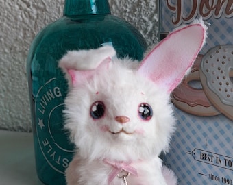 Op de afbeelding: Een wit en roze pluche konijn met grote blauwe ogen en een roze strik om zijn nek. Het konijn draagt een zilveren sleutelhanger. Het speelgoed zit op een wit oppervlak.