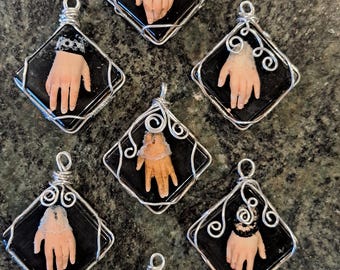 May include: Seven black square pendants with silver wire frames. Each pendant features a hand wearing a lace cuff, with the hand and cuff encased in resin.