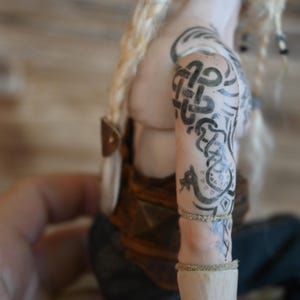 May include: A doll with blonde hair and intricate black tattoos on the arm. The tattoos feature Celtic knotwork and other designs. The doll is wearing a brown and gold outfit, with a leather strap detail.