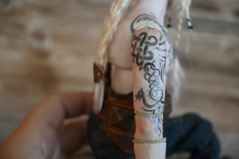 May include: A doll with blonde hair and intricate black tattoos on the arm. The tattoos feature Celtic knotwork and other designs. The doll is wearing a brown and gold outfit, with a leather strap detail.