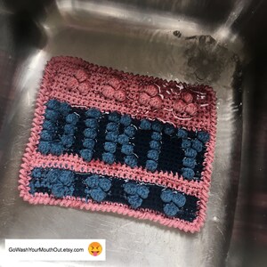 Puede incluir: Un paño de cocina de crochet rosa y azul con la palabra "sucio" escrita en hilo azul. El paño está mojado y se encuentra en un fregadero de acero inoxidable.