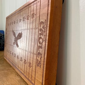 May include: A wooden cutting board with a football field design and the words "Philadelphia Eagles" engraved on it. The board is brown and has a natural wood finish.