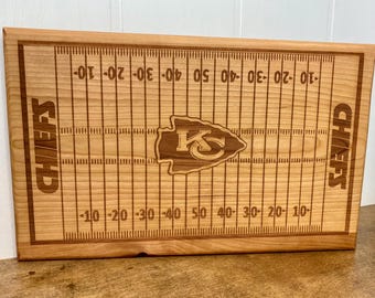 Op de afbeelding: Een houten snijplank met een Kansas City Chiefs logo en een voetbalveld ontwerp. De plank is gegraveerd met de woorden "Chiefs" aan beide kanten en heeft yard lines gemarkeerd van -50 tot 50.