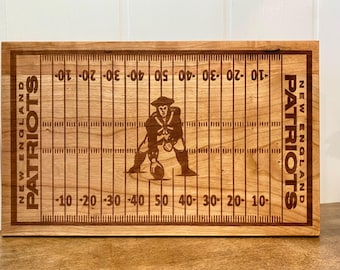 May include: A wooden chopping board with a football field design. The board features the New England Patriots logo and team name. The field lines and yard markers are etched into the wood, creating a detailed and thematic design.