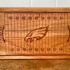 May include: A wooden cutting board with a football field design and an eagle logo. The board is engraved with the words "EAGLES" and "PHILADELPHIA".