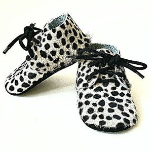 Può includere: Un paio di scarpine per neonati con stampa leopardata bianca e nera e lacci neri. Le scarpe hanno una punta arrotondata e un tacco basso.