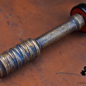May include: A close-up of a metal tool with a textured handle and a black and red cap. The tool has a bronze finish with a spiral design on the handle. The tool is on a brown leather surface.