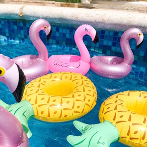 May include: A set of inflatable pool floats, including two pink flamingo drink holders, a yellow pineapple drink holder, and a green pineapple leaf. The floats are floating in a blue swimming pool.