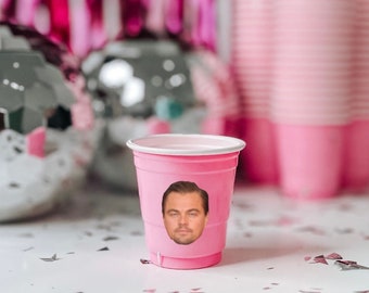 May include: A pink plastic cup with a cutout for a face. A photo of Leonardo DiCaprio's face is placed in the cutout. The cup is sitting on a white surface with silver confetti.