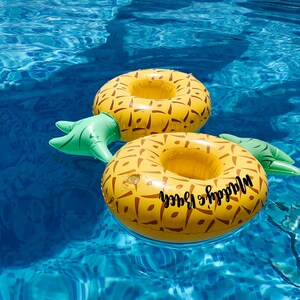 May include: Two yellow and brown inflatable pineapple-shaped drink holders floating in a blue swimming pool. Each has a green stem and the words "Maddy's Beach" in black script. The water is clear and reflects the sunlight.