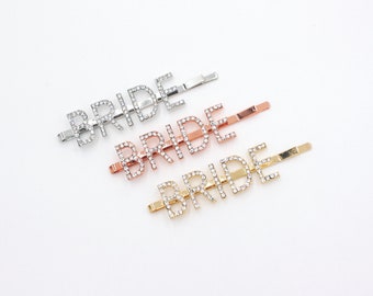 May include: Three hair barrettes spelling out the word "BRIDE" in silver, rose gold, and gold. Each barrette has the word spelled out in capital letters, with small, clear rhinestones embedded in the letters.