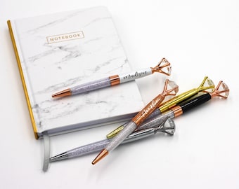 May include: A white marble notebook with gold trim and the word "NOTEBOOK" in gold lettering. Several diamond-topped pens with crystal-filled barrels are scattered around the notebook. One pen is personalized with the name "Madison".
