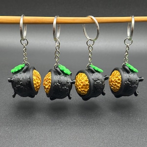 Può includere: Quattro portachiavi neri e dorati a forma di pentola con un trifoglio verde in cima. I portachiavi sono appesi a un bastoncino di legno.