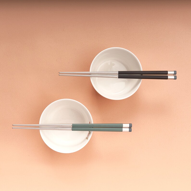 May include: Two white bowls and two pairs of chopsticks. One pair of chopsticks is black and the other pair is green.