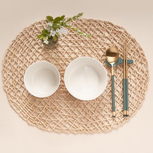 May include: Two white bowls, a gold spoon, and a pair of green chopsticks on a woven straw placemat. The bowls are empty and the spoon and chopsticks are resting on a small green holder.
